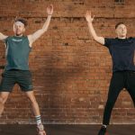 Two men performing jumping jacks in a gym setting, showcasing fitness and energy.
