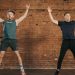 Two men performing jumping jacks in a gym setting, showcasing fitness and energy.