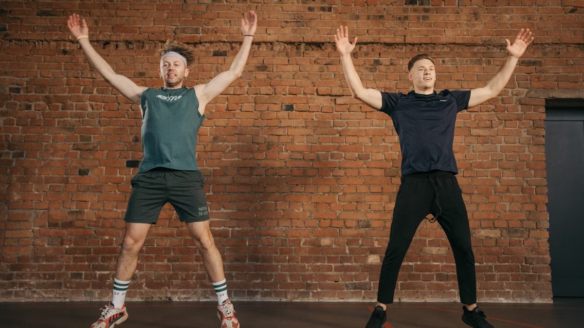 Two men performing jumping jacks in a gym setting, showcasing fitness and energy.